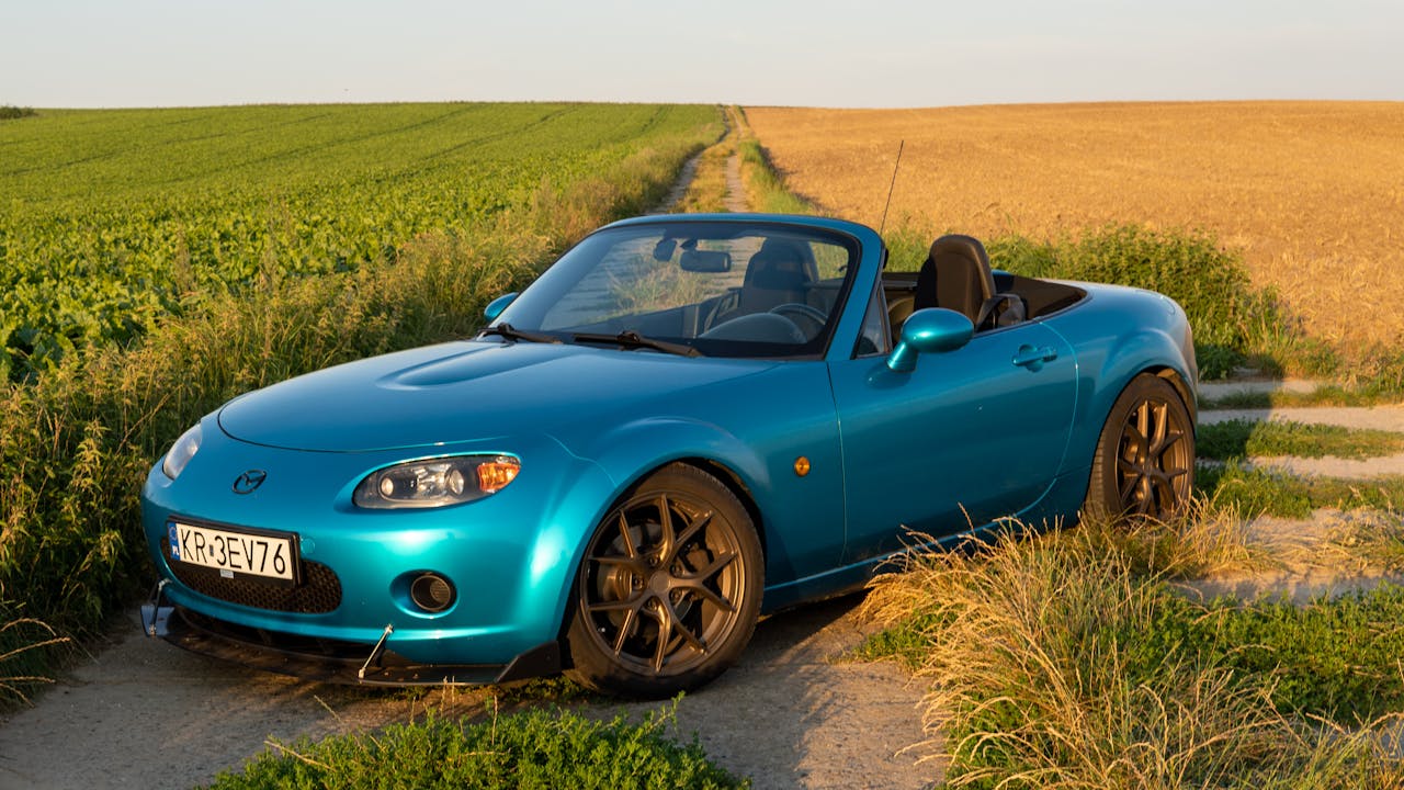 A vibrant blue Mazda MX-5 sports car on a rural dirt road in Słomniki, Poland during daylight.