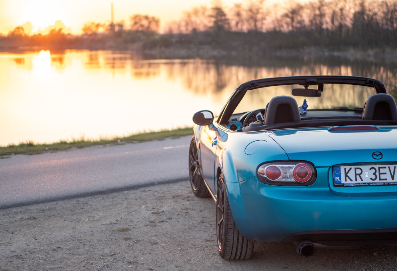 A blue convertible Mazda MX-5 parked by a lakeside road during sunset in Brzegi, Poland.